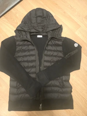 Moncler cardigan - Snygg svart pufferjacka med quiltad front och huva. Jackan har stickade ärmar och sidor, dragkedja framtill och två fickor. Moncler-logga på ärmen ger en exklusiv touch. Perfekt för kalla vinterdagar och riktigt skön att bära. För mer information är det bara att kontakta!  NFC SCAN FUNKAR!