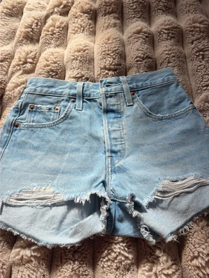 Ljusblåa högmidjade jeansshorts - Säljer ett par ljusblåa högmidjade jeansshorts med råa, fransiga kanter och slitningar framtill. Shortsen har klassisk femficksdesign, bälteshällor och knappgylf. Perfekta för varma dagar och en avslappnad look.