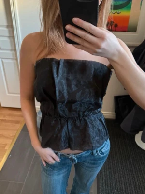 Svart  tubtopp från Gina tricot - Snygg svart axelbandslös tubtopp med lätt glansigt tyg och markerad midja. Knappt använd.
