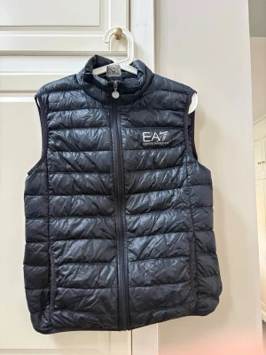 Svart dunväst från Emporio Armani - Snygg svart dunväst från Emporio Armani EA7 med quiltad design och hög krage. Västen har dragkedja framtill och logga på bröstet. Perfekt för lager-på-lager och ger en sportig vibe till din outfit.