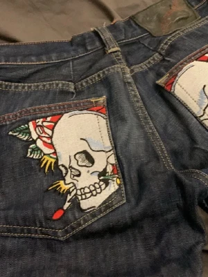 Ed Hardy Jeans  - Unika mörkblå jeans med stora broderade dödskallar och rosor på bakfickorna. Jeansen har raka ben och coola detaljer med veckade partier vid knäna. Materialet är klassisk denim i bomull. Perfekt för dig som vill sticka ut med en edgy stil.