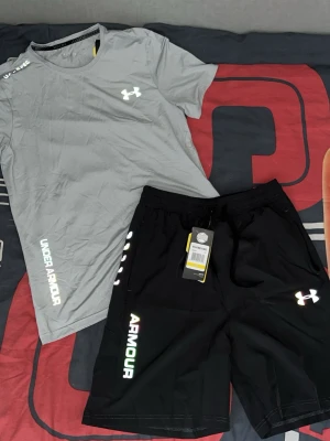 Under Armour träningsset t-shirt & shorts - Snyggt träningsset från Under Armour med en grå t-shirt och svarta shorts. T-shirten har färgglad logga på bröstet och tryck på axeln, medan shortsen har logga och text längs benet. Båda plaggen är i lätt och snabbtorkande material, perfekt för träning.