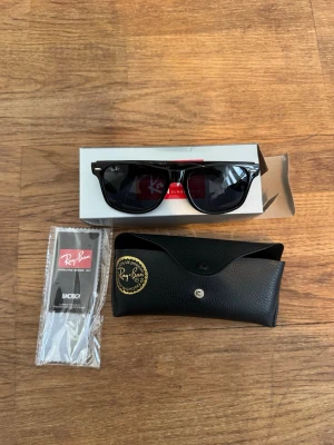 Svarta Ray-Ban Wayfarer solglasögon - Klassiska svarta Ray-Ban Wayfarer solglasögon med tidlös design. Bågarna är i blank svart plast och glasen är mörka. Kommer med originalfodral och putsduk. Perfekta för att höja din stil på sommaren.