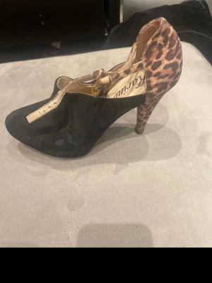 Svarta pumps med leopardmönster - Unika pumps med svart mockaliknande material framtill och leopardmönstrad klack och häl. Skon har en T-rem i leopardmönster som ger extra stöd och en cool kontrast. Perfekta för dig som vill sticka ut med din stil.
