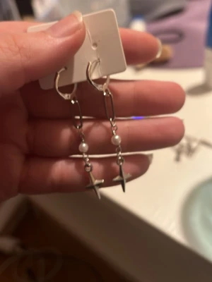 Silvriga örhängen med kors och pärlor - Säljer ett par trendiga silvriga örhängen med hängande kors och små vita pärlor. Örhängena har en cool kedjedesign och passar perfekt för dig som gillar edgy accessoarer med en twist. Snygga att matcha med flera örhängen eller bära som statement.