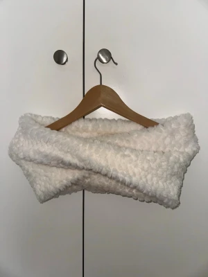 Vit fluffig tubhalsduk - Mysig och supermjuk tubhalsduk i fluffigt vitt material. Halsduken har en twistad design och känns riktigt varm och skön. Perfekt för kalla dagar när du vill ha något gosigt runt halsen.