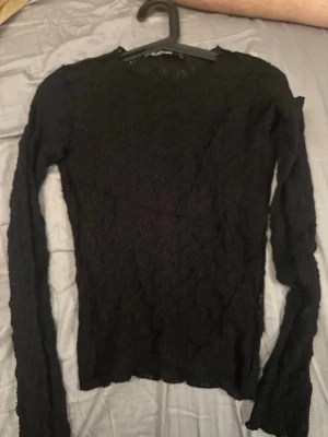 Svart spetsblus med lång ärm - Säljer en svart, långärmad blus i spets med rund hals. Toppen har en figurnära passform och är helt täckt av ett fint spetsmönster som ger en elegant look. Perfekt att styla med jeans eller kjol för en trendig outfit.