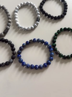 Set med pärlarmband i olika färger - Säljer ett set med fem elastiska armband gjorda av runda pärlor i olika färger: blå, grön, vit, svart och grå. Varje armband har en unik färgkombination och vissa har inslag av metalliska detaljer. Perfekt för att mixa och matcha till din stil.