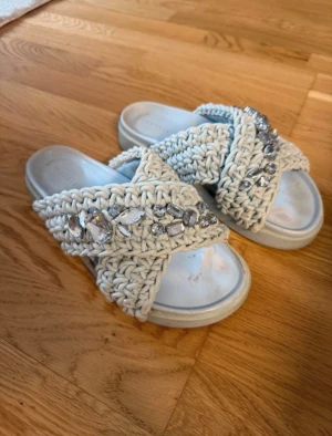 Inuikii tofflor  - Säljer ett par ljusblå sandaler med breda, stickade band i kors och stora dekorativa stenar på ovansidan. Sulan är platt och bekväm, och banden har en chunky virkad look som ger en trendig vibe. Perfekta för dig som vill ha något unikt och snyggt till sommaren.