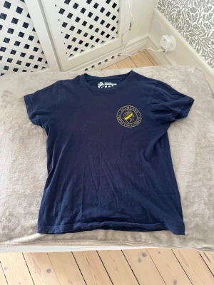 Mörkblå AIK t-shirt med tryck - Snygg mörkblå t-shirt med AIK:s klassiska klubbmärke tryckt på bröstet. T-shirten har rund hals och korta ärmar, perfekt för dig som vill visa ditt stöd för laget. Tillverkad i mjuk bomull för skön känsla hela dagen.