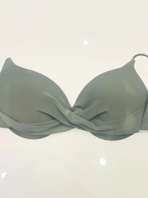 Mörk grön bikini överdel med twist framtill - Stilren mörkgrön bikini med vadderade kupor och bygel. Framtill har den en snygg twistdetalj som ger en modern look. Axelbanden är justerbara och materialet känns mjukt och följsamt mot huden. Perfekt för dig som vill ha en enkel men trendig bikinitopp.