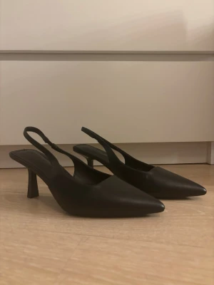 Svarta slingback pumps med spetsig tå - Stilrena svarta pumps med spetsig tå och smal klack. Skorna har en öppen häl med justerbar slingback-rem. Perfekta för att lyfta vilken outfit som helst. Aldrig använda och har orginal boxen med som man får dem skickade i! 