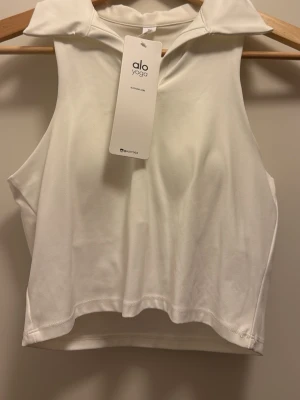 Vit ärmlös topp från Alo Yoga - Snygg vit ärmlös topp från Alo Yoga med krage och croppad passform. Perfekt för dig som gillar sportig och stilren look. Tillverkad i mjukt och stretchigt material som känns skönt mot huden.