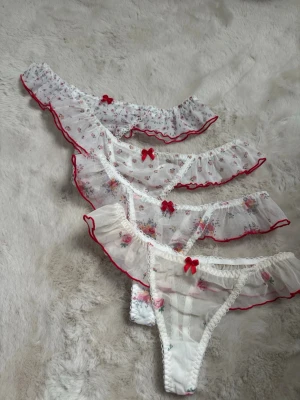 Söta vita spetsiga trosor med volang - Fyra par vita trosor i transparent mesh med volangkant och små blommiga mönster. Varje par har en röd rosett framtill och röda detaljer längs kanterna. Romantisk och feminin design, perfekt för dig som gillar gulliga underkläder.