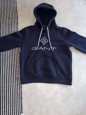 Mörkblå hoodie från GANT - Snygg mörkblå hoodie från GANT med vit logga och tryck framtill. Klassisk huva med vita dragsnören och en stor känguruficka. Perfekt för en avslappnad och stilren look. Mjuk bomullsblandning som känns skön mot huden.