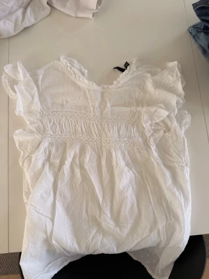 Vit blus med volanger och broderi - Söt vit blus med volangdetaljer vid ärmar och hals, samt smockad midja och diskret broderat mönster. Blusen har en lätt v-ringning och är tillverkad i ett tunt, luftigt tyg som känns perfekt för sommaren.