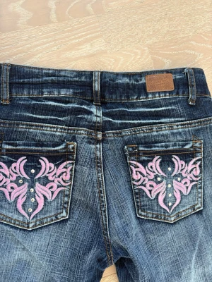 Chilly girl bootcut jeans - Snygga blå jeans med slitningar och rosa broderade detaljer på bakfickorna samt rosa sömmar fram. Jeansen har normal midja och raka ben. Materialet är klassisk denim med cool tvätt och dekorativa nitar på fickorna. Perfekta för en trendig och unik stil.