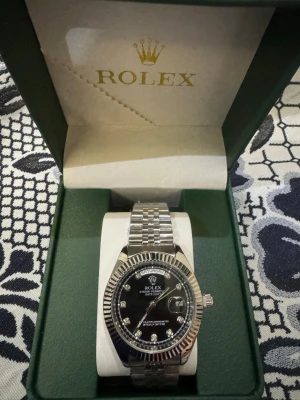 R silverklocka med svart urtavla - Snygg R klocka i silver med länkarmband och svart urtavla. Urtavlan har datumvisning, veckodag och markerade index med stenar. Klassisk räfflad bezel och elegant design. Perfekt accessoar för dig som gillar exklusiva detaljer. Med plast runt klockan. Funkar på batteri 