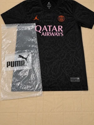 Svart PSG matchtröja - Snygg svart Paris Saint-Germain matchtröja med diskret mönster, orange PSG-logga och Jordan-logga på bröstet. Tryck i lila med 'Qatar Airways' på framsidan. Tillverkad i lätt och ventilerande material, perfekt för fotboll eller träning.