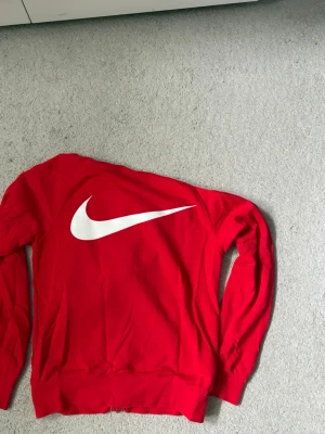 Röd Nike hoodie med Swoosh-tryck - Röd hoodie från Nike med stor vit Swoosh-logga på ryggen och svart text 'SWOOSH' framtill. Dragkedja och två mindre vita Swoosh-loggor på bröstet. Perfekt för en sportig och avslappnad stil.