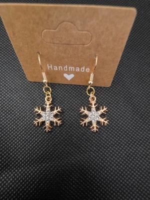 Guldiga snöflingeörhängen - Unika handgjorda örhängen i form av snöflingor i guldig metall med glittrande detaljer i mitten. Perfekta för dig som vill ha en vinterinspirerad accessoar med lite extra bling. Hängande modell med krok.