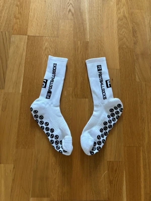 Vita FS Footballsocks med grepp - Säljer ett par vita FS Footballsocks med svart text längs sidan och greppande prickar under sulan. Perfekta för fotboll eller annan sport där du vill ha extra fäste i skorna. Klassisk ribbad design och sportig look.