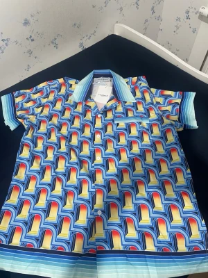 Färgglatt set med retro mönster - Säljer ett set med skjorta och shorts i ett färgstarkt retroinspirerat mönster i blått, gult, rött och vitt. Skjortan har kort ärm, krage och knappar framtill, medan shortsen har resår i midjan och dragsko. Båda plaggen har matchande grafiskt tryck och blå/vit randig kant.Vi kan komma överens med priset