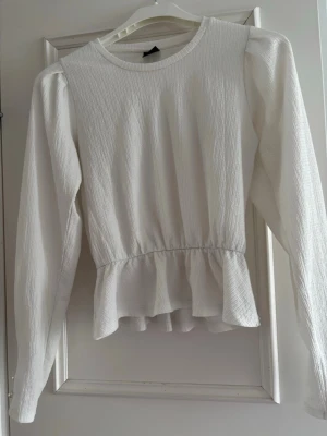 Vit långärmad blus med volang - Säljer en stilren vit blus med lång ärm och rund halsringning. Blusen har en markerad midja och volang nedtill som ger en feminin siluett. Materialet är lätt strukturerat och ger en snygg textur. Perfekt att matcha med jeans eller kjol.