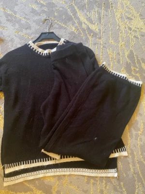 Svart stickat loungewear-set - Mysigt svart loungewear-set med tröja och byxor. Båda plaggen har vita ribbade detaljer vid hals, ärmslut, benslut och nederkant. Setet är stickat och har en avslappnad passform, perfekt för chill dagar hemma.