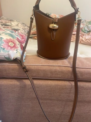 Viral väska H&M - Säljer den här snygga bruna virala bucket axelväska med justerbar rem och guldfärgat spänne. Väskan har en unik cylinderform och ett stilrent handtag upptill. Perfekt för dig som vill ha en trendig och praktisk accessoar till vardagslooken. Använd endast fåtal gånger, så det finns inga tecken på använding