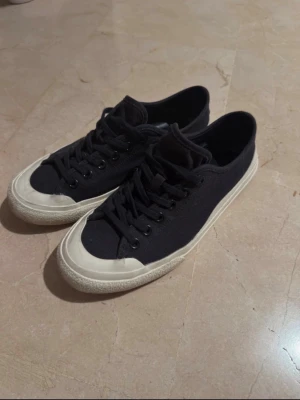 Svarta låga sneakers med vit sula - Snygga svarta sneakers med lågt skaft och klassisk vit gummisula. Skorna har svart tygöverdel, svarta skosnören och tåhätta i vitt gummi. Perfekta för en avslappnad och stilren look.