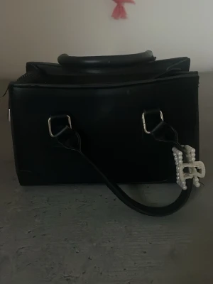 Svart handväska med pärldetalj - Snygg svart handväska i boxig modell med två handtag och silverfärgade metalldetaljer. Väskan har en dekorativ hårklämma som ingår på ena handtaget. Perfekt för dig som gillar stilrena accessoarer med en twist.