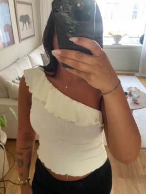 Vit oneshoulder topp med volang - Säljer en superfin vit oneshoulder topp med volangdetalj längs halsringningen. Toppen är croppad och har en lätt ribbad struktur som ger en snygg passform. Perfekt till jeans eller kjol för en trendig look.