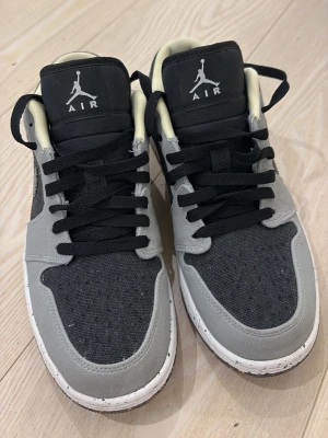 Grå och svarta Air Jordan 1 Low - Snygga Air Jordan 1 Low sneakers i grått och svart med svart snörning och vit sula med svarta prickar. Ovandelen är i textil och mocka, med blå detaljer på hälen och klassisk Jumpman-logga på plösen. Perfekta för dig som gillar streetwear.