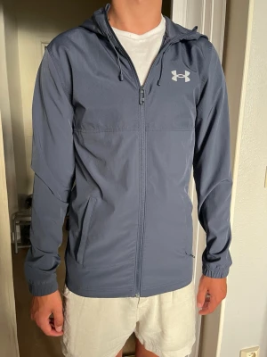 Blå vindjacka från Under Armour - Snygg blå vindjacka från Under Armour med huva och dragkedja framtill. Jackan har två sidofickor, resår vid ärmsluten och logga på bröstet samt stor text på ryggen. Tillverkad i lätt och tunt material, perfekt för blåsiga dagar.