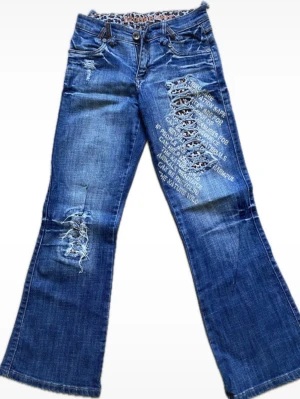 Blå jeans med snörning och tryck - Unika blå jeans med vida ben, snörningsdetaljer och tryck på både fram- och baksida. Jeansen har slitningar och broderade texter som ger en cool streetwear-vibe. Tillverkade i klassisk denim med låg midja och fem fickor.