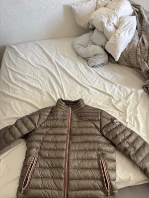 Beige dunjacka med randiga detaljer - Snygg beige dunjacka med horisontella sömmar och två dragkedjefickor med randiga detaljer. Jackan har hög krage och en diskret logotyp på ärmen. Perfekt för kalla vinterdagar och riktigt lätt att bära.