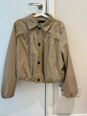  Ny trendig Beige kort jacka med puffärm - Snygg beige jacka med puffiga ärmar och resår i midjan. Jackan har klassisk krage, knäppning framtill med svarta knappar och två fickor. Tillverkad i ett mjukt bomullsliknande material och har en avslappnad, boxig passform.