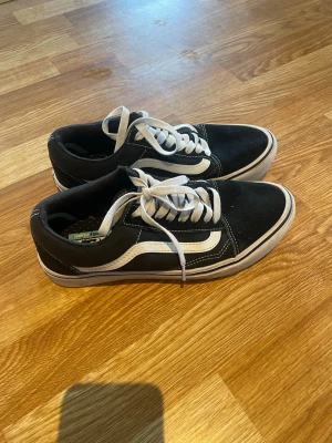 Svarta Vans Old Skool sneakers - Klassiska Vans Old Skool sneakers i svart med vit sula och vit sidostrip. Skorna har vita skosnören och är tillverkade i mocka och canvas. Perfekta för en avslappnad streetstil och passar till det mesta. 