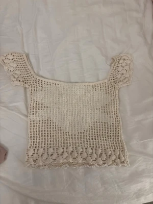 Virkat offshoulder topp i beige - Superfin virkad topp i beige med offshoulder-modell och korta ärmar. Toppen har ett luftigt, rutigt mönster och dekorativa virkade detaljer längs nederkanten och ärmarna. Perfekt för sommarens alla tillfällen!
