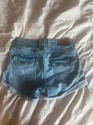Blå denimshorts med uppvik - Snygga blå jeansshorts i klassisk modell med uppvikta benslut och fyra fickor. Shortsen har bälteshällor och knappgylf. Perfekta för varma dagar och enkla att matcha med olika stilar.