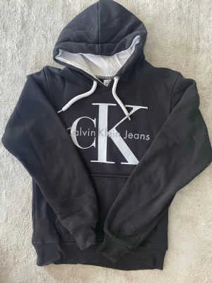 Svart tröja CK - Svart hoodie från Calvin Klein Jeans med stor vit CK-logga och text på bröstet. Huvan har vit insida och vita dragsnören. Tyget är tjockt och varmt den passar även XS-M 