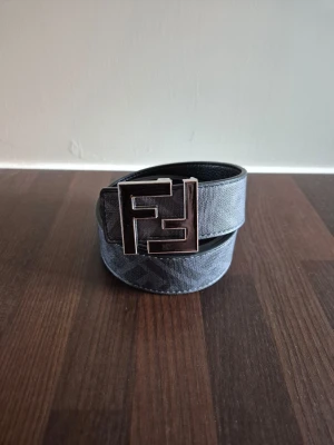 FF bälte - Snyggt bälte från Fendi i svart och grått med ett stort metallspänne med Fendi-logga. Bältet har diskret mönster och är tillverkat i skinn med textil på utsidan. Perfekt accessoar för att lyfta din outfit.