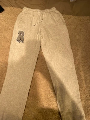 Grå mjukisbyxor med broderad figur - Snygga ljusgrå mjukisbyxor med resår och dragsko i midjan. På vänster lår finns en broderad tecknad figur i grått och svart. Byxorna har raka ben och är gjorda i mjukt bomullsmaterial. Perfekta för chill dagar hemma eller när du vill vara bekväm.