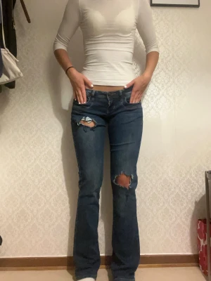 Mörkblå bootcut jeans med slitningar - Snygga mörkblå bootcut jeans med slitningar på båda benen och klassiska fem fickor. Jeansen har låg midja och detaljerade bakfickor med dekorativa sömmar. Perfekta för en avslappnad och trendig look.