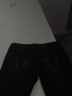Svarta bootcut lågmidjade jeans med detaljer där bak  - Snygga svarta bootcut jeans med klassisk femficksmodell och silverfärgade detajler på bakfickorna. Jeansen har låg midja och stängs med knapp och dragkedja. Perfekta för dig som gillar låg midjad stil. 