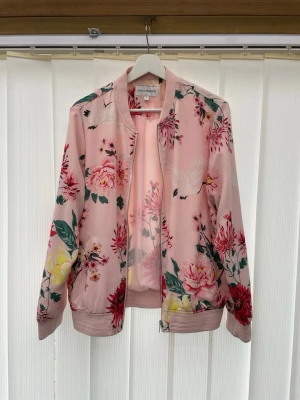 Rosa bomberjacka med blommigt mönster - Snygg rosa bomberjacka från KappAhl i storlek M med blommigt mönster. Jackan har ribbade muddar och krage, dragkedja framtill och är tillverkad i ett lätt, glansigt material. Perfekt för dig som vill sticka ut med en färgglad och trendig look.
