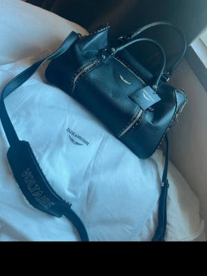 Svart handväska Zadig & Voltaire Sunny Medium size - Cool svart handväska i läder från Zadig & Voltaire med silverfärgade nitar och detaljer. Väskan har dubbla handtag, avtagbar axelrem med logga och dragkedjor framtill. Perfekt för dig som gillar edgy och trendig stil.