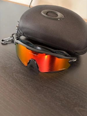 Oakley sportiga solglasögon svart/röd - Säljer ett par sportiga Oakley solglasögon med svart båge och spegelröda linser. Modellen har en halvbåge och sitter skönt på ansiktet, perfekt för dig som gillar en aktiv livsstil. Kommer med ett hårt skyddsfodral med Oakley-logga.