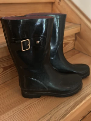 Svarta gummistövlar med spänne - Snygga svarta gummistövlar med dekorativt spänne i metall på sidan. Stövlarna har en klassisk form och är helsvarta med en något högre sula. Perfekta för regniga dagar och festivaler.